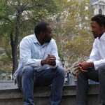 Two black men having a conversation while sitting on a stone wall